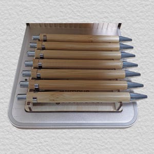 May include: A set of seven retractable pens with light brown bamboo barrels and silver-toned metal clips, tips, and push-button mechanisms. The pens are arranged horizontally and stacked in a light-colored wooden display stand with a grooved base.