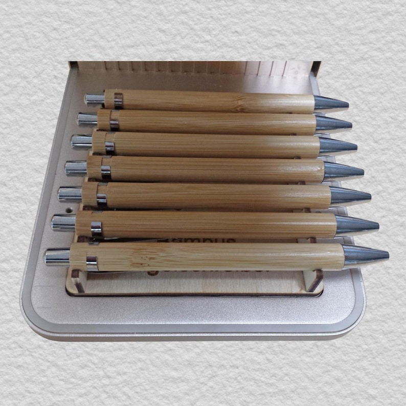 May include: A set of seven retractable pens with light brown bamboo barrels and silver-toned metal clips, tips, and push-button mechanisms. The pens are arranged horizontally and stacked in a light-colored wooden display stand with a grooved base.