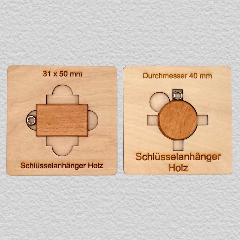 May include: Two light-colored wooden keychains set in square templates. The left rectangular piece measures 31 x 50 mm, and the right circular piece has a diameter of 40 mm. Both pieces feature a small metal ring and the text Schl&uuml;sselah&auml;nger Holz.
