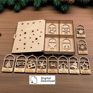 Könnte beinhalten: Ein Set aus hellen Holz-Weihnachtsanhängern mit verschiedenen ausgeschnittenen Motiven wie Sternen, Glocken, Schneeflocken und Rentieren, aufbewahrt in einer passenden Holzbox mit Sternenmuster-Deckel. Jeder Anhänger trägt den Text Frohe Weihnachten.