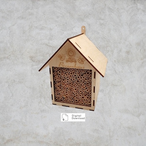 Può includere: Una casetta per api in legno marrone chiaro con tetto a capanna e apertura rettangolare riempita di tubi di bambù. La casetta presenta la scritta "Bienenhaus" e illustrazioni di api e fiori. Un piccolo anello in alto.