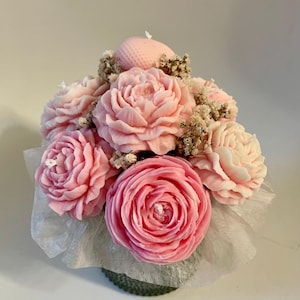 Könnte beinhalten: Ein Arrangement aus rosa pfingstrosenförmigen Kerzen mit einer kleinen, runden Kerze obenauf. Die Kerzen sind in einem kleinen, grünen Behälter angeordnet, mit weißem Seidenpapier und Trockenblumen. Die Kerzen sind hellrosa.