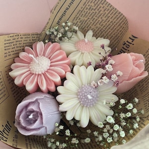 Könnte beinhalten: Ein Bouquet aus blütenförmigen Kerzen in Rosa-, Weiß- und Lavendeltönen, verpackt in Vintage-Zeitungspapier. Die Kerzen sind detailliert, um Gänseblümchen, Rosen und anderen Blüten zu ähneln, mit kleinen weißen Blüten dazwischen.