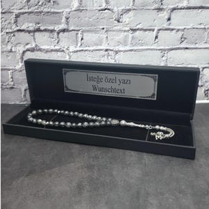 Pode incluir: Um terço com contas de pedra cinza-escura manchadas e detalhes em metal prateado, incluindo uma borla decorativa e um pingente de letra. O terço é apresentado numa caixa retangular preta com o texto "İsteğe özel yazı" e "Wunschtext" impresso na tampa interior.