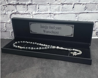 Op de afbeelding: Een gebedsketting met donkergrijze, gemarmerde stenen kralen en zilverkleurige metalen accenten, inclusief een decoratief kwastje en een letterbedel. De ketting wordt gepresenteerd in een zwarte rechthoekige doos met de tekst "İsteğe özel yazı" en "Wunschtext" op de binnenkant van het deksel.