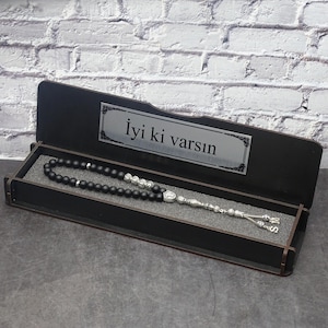 May include: A black beaded prayer bead set with silver accents, presented in an open black wooden box. The box has a silver plaque with text. The beads are arranged on a gray foam insert.