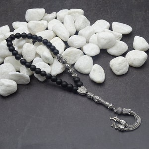 May include: A black beaded prayer bead necklace with silver-colored decorative elements. The beads are matte and round. The silver pieces have intricate patterns and a tassel at the end.
