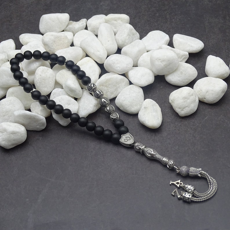 May include: A black beaded prayer bead necklace with silver-colored decorative elements. The beads are matte and round. The silver pieces have intricate patterns and a tassel at the end.
