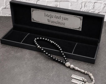 May include: A string of black prayer beads with silver-toned metal accents and two rectangular silver-toned tags engraved with names, presented in a black rectangular gift box featuring a silver plaque with the text Isteğe özel yazı Wunschtext.