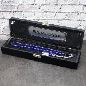 Peut inclure: Un chapelet de perles de prière bleu translucide avec des accents en métal argenté, présenté dans un coffret rectangulaire doublé de velours noir. Le couvercle intérieur comporte une plaque en métal argenté gravée Baba, seni seviyoruz.