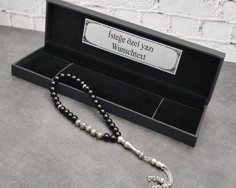May include: A string of black prayer beads with silver-toned accents and a decorative silver-toned tassel, presented in a black rectangular box. The box lid features a white label with the text "İsteğe özel yazı" and "Wunschtext" printed on it.