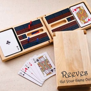 May include: A wooden cribbage board game with a folding design. The board has a red and blue playing surface with a wooden box for storing the pegs. The box has a wooden lid with the text "Reeves Get Your Game On!" engraved on it.