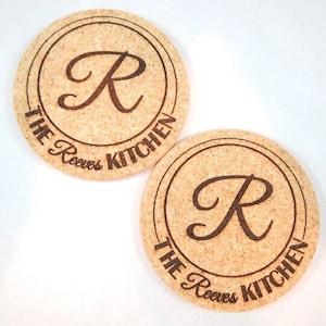 May include: Two round cork coasters with a brown woodgrain finish. Each coaster features a circle with the letter "R" in a cursive font and the text "The Reeves Kitchen" around the circle.