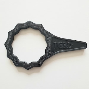 May include: Black 3D-printed tool with a circular, gear-like head and a handle. The handle has the text "TG3D" printed on it. The tool is made of a matte black material and is set against a white background.