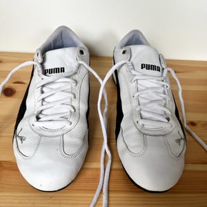 Pu&ograve; includere: Un paio di scarpe da ginnastica Puma in pelle bianca con dettagli neri e lacci bianchi. Il logo Puma &egrave; visibile sul lato di ogni scarpa.