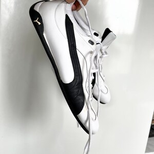 Pu&ograve; includere: Un paio di scarpe da ginnastica in pelle bianca con dettagli e lacci neri. Le scarpe hanno un design a collo basso e una suola in gomma.