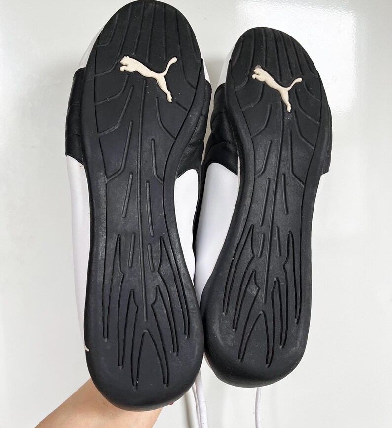 Pu&ograve; includere: Un paio di scarpe da ginnastica Puma bianche e nere con suola testurizzata. Le scarpe hanno una tomaia in pelle bianca e una suola in gomma nera.