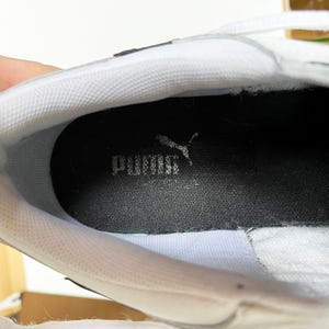 Pu&ograve; includere: Primo piano dell'interno di una scarpa da ginnastica bianca con una soletta interna nera su cui &egrave; scritto "PUMA" in lettere bianche.