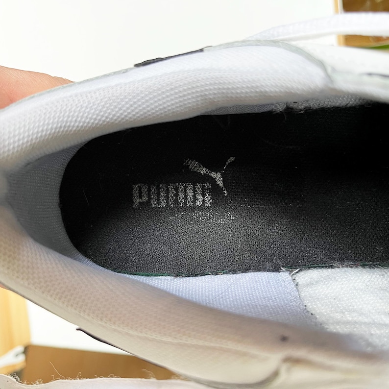 Pu&ograve; includere: Primo piano dell'interno di una scarpa da ginnastica bianca con una soletta interna nera su cui &egrave; scritto "PUMA" in lettere bianche.