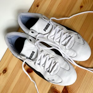 Pu&ograve; includere: Un paio di scarpe da ginnastica Puma in pelle bianca con dettagli neri e lacci bianchi. Il logo Puma &egrave; visibile sul lato di ogni scarpa.