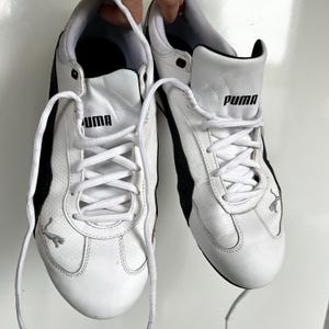 Pu&ograve; includere: Un paio di scarpe da ginnastica Puma in pelle bianca con dettagli e lacci neri. Il logo Puma &egrave; visibile sul lato di ogni scarpa.