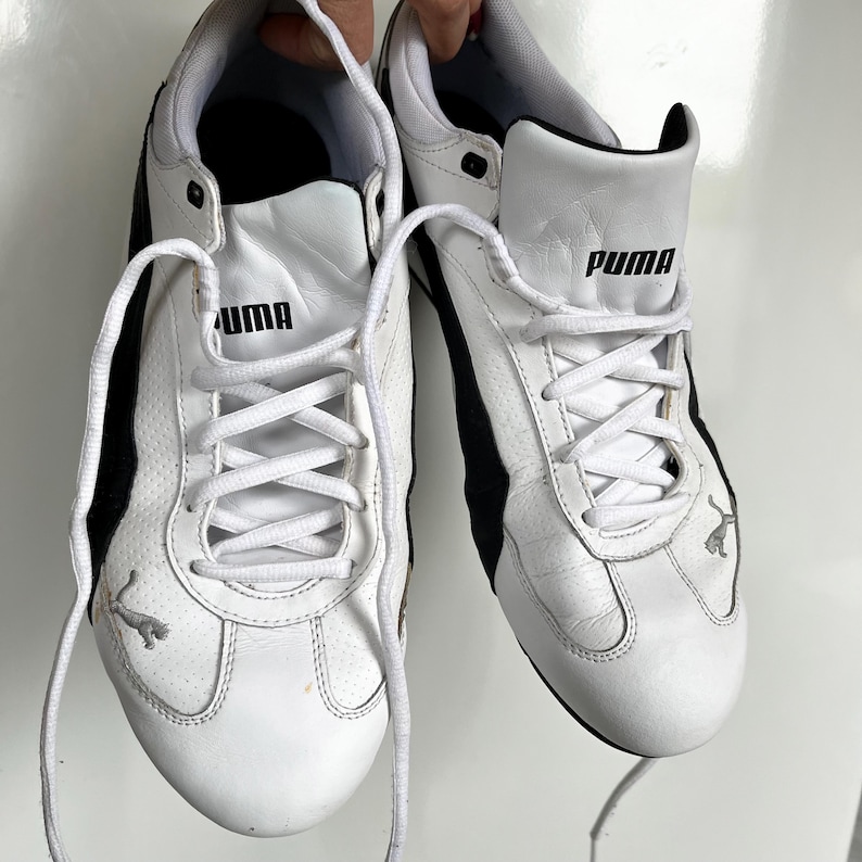 Pu&ograve; includere: Un paio di scarpe da ginnastica Puma in pelle bianca con dettagli e lacci neri. Il logo Puma &egrave; visibile sul lato di ogni scarpa.