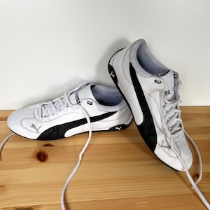 Pu&ograve; includere: Un paio di scarpe da ginnastica Puma in pelle bianca con dettagli neri. Le scarpe hanno un logo Puma nero sul lato e una striscia nera sul retro.
