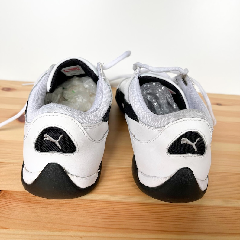 Pu&ograve; includere: Un paio di scarpe da ginnastica Puma in pelle bianca con dettagli neri. Le scarpe hanno un logo Puma nero sul retro e una suola in gomma nera.