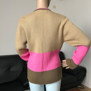 May include: A long-sleeved, ribbed knit sweater featuring a color-blocked design. The upper portion is tan, followed by a wide horizontal band of bright pink across the midsection, and a dark brown hem. The collar is also trimmed in dark brown.