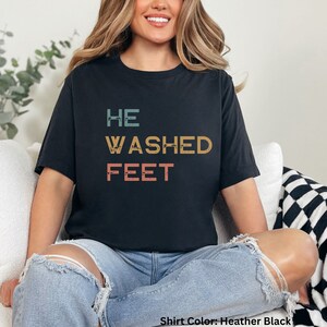 May include: A black t-shirt with the text "HE WASHED FEET" in a vintage style font. The text is in shades of blue, orange, and pink.