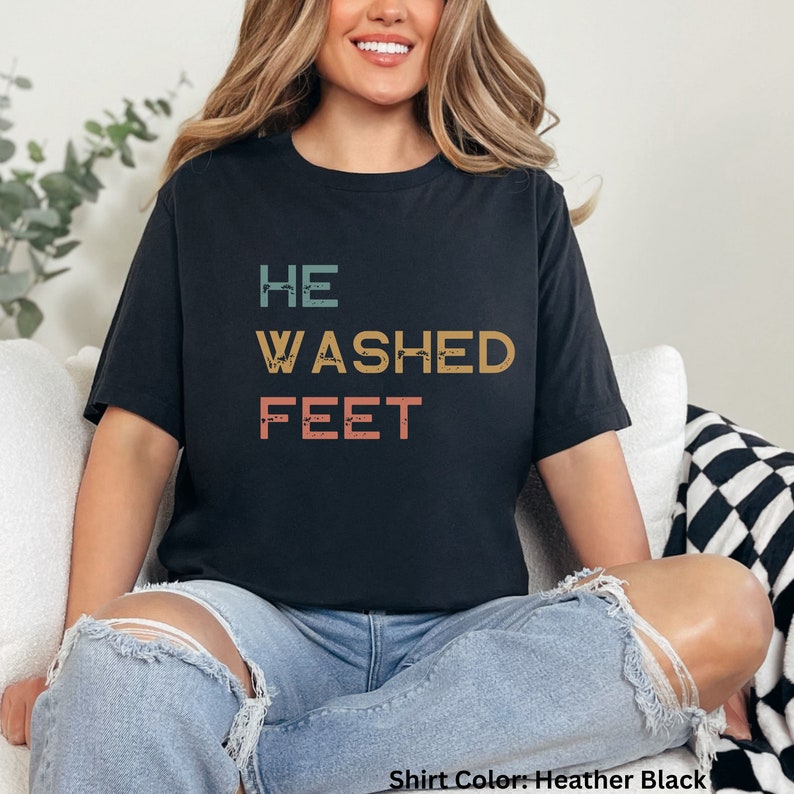 May include: A black t-shirt with the text "HE WASHED FEET" in a vintage style font. The text is in shades of blue, orange, and pink.
