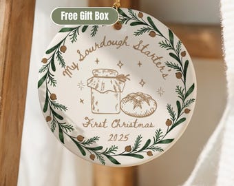 May include: A round, off-white ceramic ornament with a green and brown botanical border. The ornament features the text "My Sourdough Starter's First Christmas 2025" and illustrations of a jar and a loaf of bread.