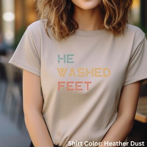 May include: A heather dust colored t-shirt with the text "HE WASHED FEET" in a vintage style font with colorful letters.