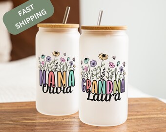 May include: Two frosted glass tumblers with bamboo lids and metal straws. Each tumbler features a floral design with the words "NANA Olivia" and "GRANDMA Laura" in colorful letters. The tumblers are on a wooden surface.