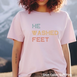 May include: A soft pink t-shirt with the text "HE WASHED FEET" in a vintage style font. The text is in shades of blue, green, orange, and yellow.