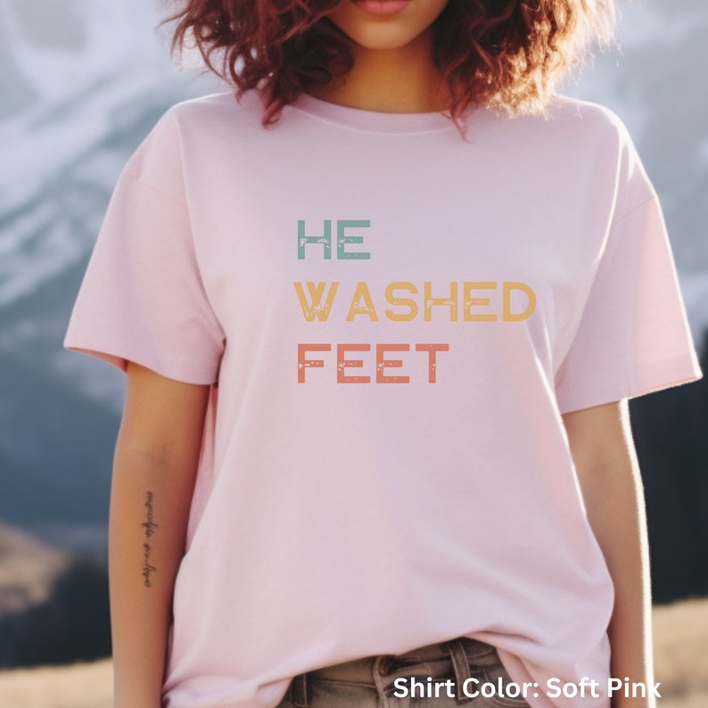 May include: A soft pink t-shirt with the text "HE WASHED FEET" in a vintage style font. The text is in shades of blue, green, orange, and yellow.