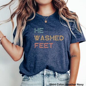 May include: A heather navy blue t-shirt with the text "HE WASHED FEET" in a vintage style font with colorful letters.