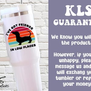 May include: White tumbler with a handle featuring a dachshund silhouette and the text "I'VE GOT FRIENDS IN LOW PLACES" above a colorful sunset design. The tumbler also has the text "KLS Guarantee".