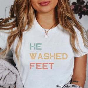 May include: White t-shirt with the text "HE WASHED FEET" in a vintage style font with faded colors. The text is in a vertical stack with each word in a different color.