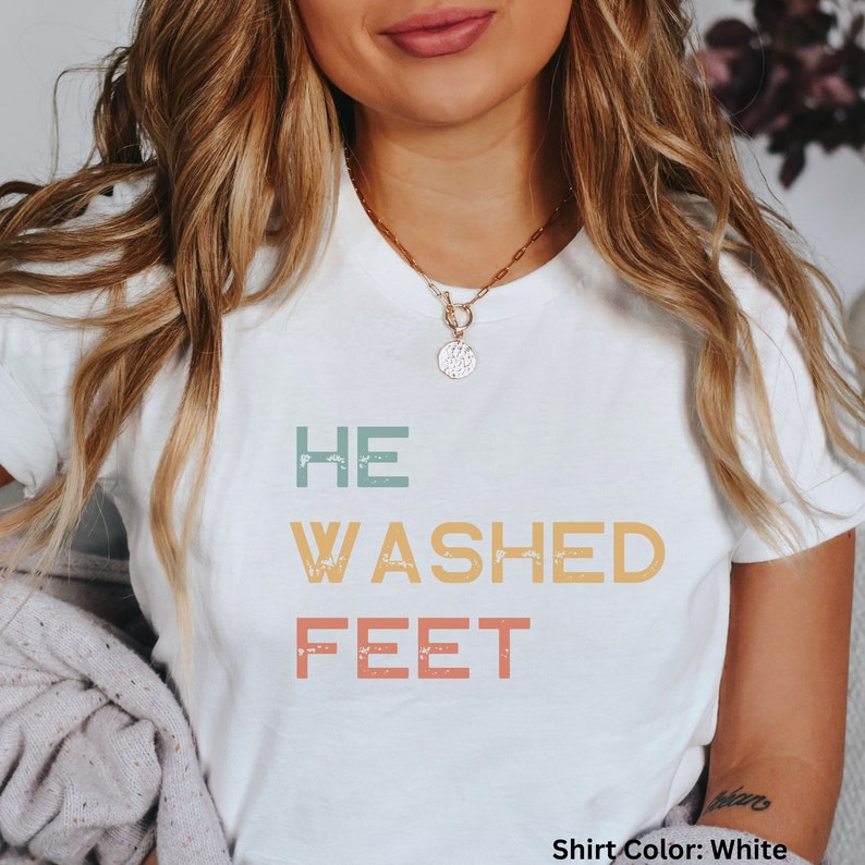 May include: White t-shirt with the text "HE WASHED FEET" in a vintage style font with faded colors. The text is in a vertical stack with each word in a different color.