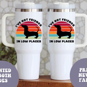May include: Two white travel mugs with a handle, featuring a black dachshund silhouette against a sunset design. The text reads "I'VE GOT FRIENDS IN LOW PLACES." Additional text states "PRINTED ON BOTH SIDES" and "PRINT NEVER FADES."