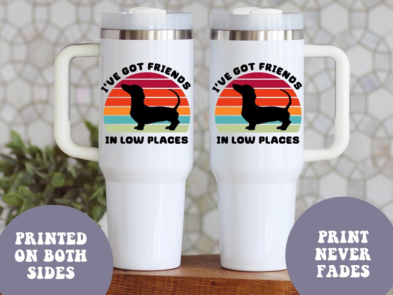May include: Two white travel mugs with a handle, featuring a black dachshund silhouette against a sunset design. The text reads "I'VE GOT FRIENDS IN LOW PLACES." Additional text states "PRINTED ON BOTH SIDES" and "PRINT NEVER FADES."