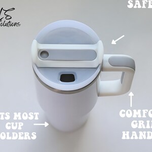 May include: White travel mug with a gray lid and handle. Features include a comfort grip handle, and the text "FITS MOST CUP HOLDERS" and "SAFE". The mug is designed for easy use and portability.