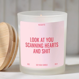 May include: A frosted glass soy wax candle with a light pink label featuring the text KLSGIFTS, LOOK AT YOU SCANNING HEARTS AND SHIT, 8OZ, and 35 H. The candle has a separate round bamboo lid with a visible wood grain texture placed to the left.