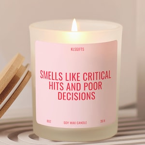 May include: A frosted glass soy wax candle with a light pink label featuring the text KLSGIFTS, SMELLS LIKE CRITICAL HITS AND POOR DECISIONS, 8OZ, and 35 H. The candle has a lit wick and is accompanied by a separate circular bamboo lid.