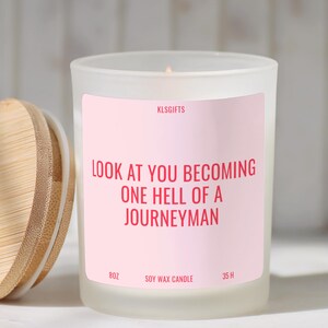 May include: A frosted glass candle in a jar with a light pink label featuring the text KLSGIFTS, LOOK AT YOU BECOMING ONE HELL OF A JOURNEYMAN, 8OZ, SOY WAX CANDLE, 35 H. The candle has a round bamboo lid placed to the left of the glass container.