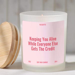 May include: A frosted glass candle jar with a light pink label featuring the text KLSGIFTS, Keeping You Alive While Everyone Else Gets The Credit, 8oz, Soy Wax Candle, and 35 H. The candle has a round bamboo lid with a white silicone seal.