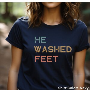 May include: A navy blue t-shirt with the text "HE WASHED FEET" in a vintage style font with a distressed effect. The text is in shades of teal, orange, and pink.