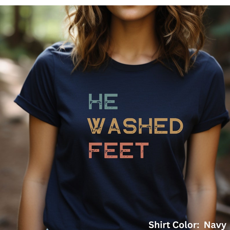 May include: A navy blue t-shirt with the text "HE WASHED FEET" in a vintage style font with a distressed effect. The text is in shades of teal, orange, and pink.