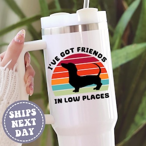 May include: White travel mug with a handle, featuring a black dachshund silhouette against a colorful sunset design. The text "I'VE GOT FRIENDS IN LOW PLACES" is printed above and below the dog.